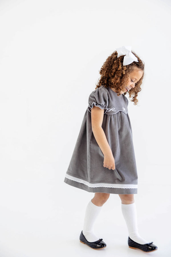 A young girl with curly hair and a white bow wears The Beaufort Bonnet Company’s McFarland Frock in Grantley Gray Corduroy with Worth Avenue White, paired with white knee socks and black shoes. She is looking down while walking against a plain white background.