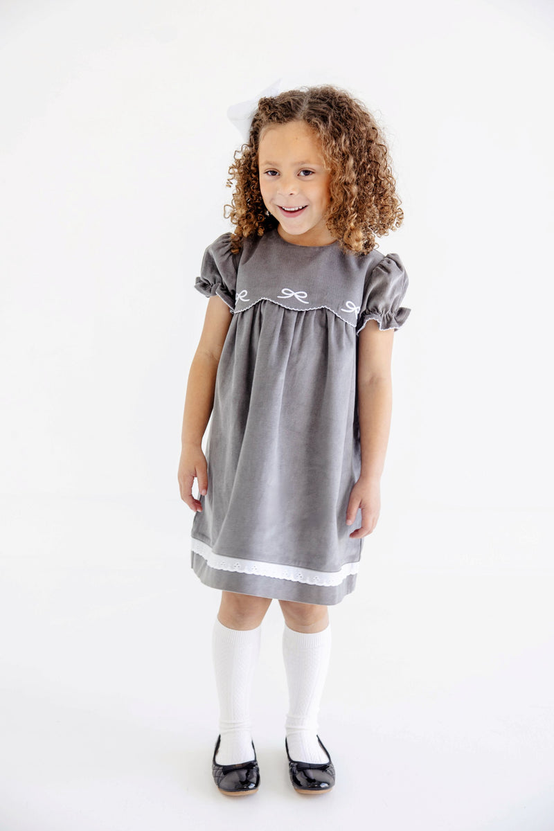 A young girl with curly hair, wearing The Beaufort Bonnet Company’s McFarland Frock in Grantley Gray Corduroy with Worth Avenue White, white knee-high socks, and black shoes, stands smiling against a plain white background.