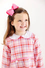 A young girl with freckles wears a long-sleeved Lindy’s Lunch Dress - Broadway Play Plaid with Worth Avenue White, featuring a white collar and a large pink hair bow.