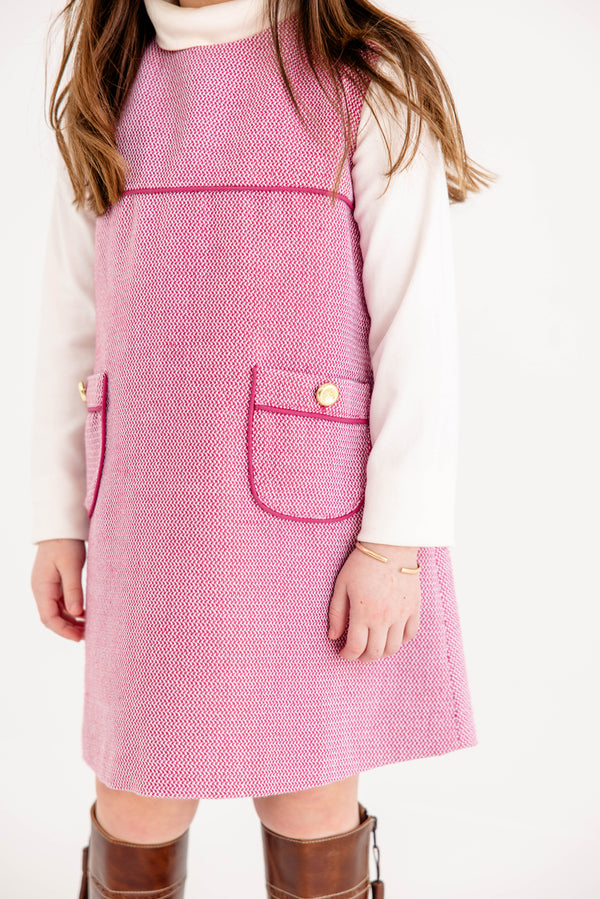 A child is wearing The Beaufort Bonnet Company’s Jill Jumper in Begley Berry Tweed over a white turtleneck shirt, paired with brown knee-high boots and a simple bracelet, standing against a white background.