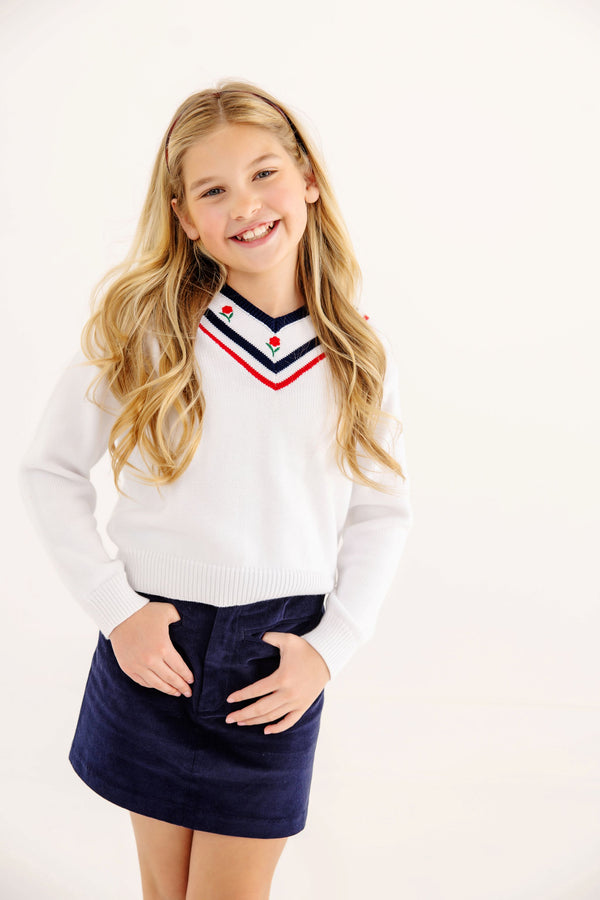 A girl wearing a Vivie June V-Neck Sweater - Worth Avenue White with Richmond Red & Nantucket Navy & Flowers, featuring a striped neckline, red and navy trim, small rose embroidery, and a navy skirt.