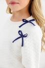 A girl with long blonde hair wears The Beaufort Bonnet Company’s Cassidy Comfy Crewneck in Quilted Palmetto Pearl, featuring two Nantucket Navy bows on the shoulder.