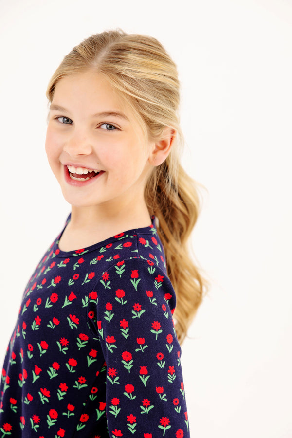 A young girl smiling in a Sadie Sweatshirt Dress - Prescott Farm Flowers with Worth Avenue White, featuring a navy blue base and a red floral print.