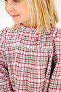 A smiling young child with blonde hair wears The Beaufort Bonnet Company’s Long Sleeve Mary Baker Day Dress in Wicker Park Plaid with Richmond Red—featuring three red buttons on the chest, it’s the perfect Christmas holiday dress.