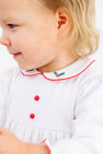A toddler wearing a Long Sleeve Britt Romper - Worth Avenue White with Holly, a white Pima cotton romper featuring red buttons and a rounded collar embroidered with holly leaves and red berries.