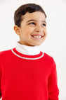 A young boy smiles while wearing an Isaac’s Intarsia Sweater - Richmond Red with Grantley Gray and Dogs, a red cotton top with a white mock neck and grey trim at the collar.