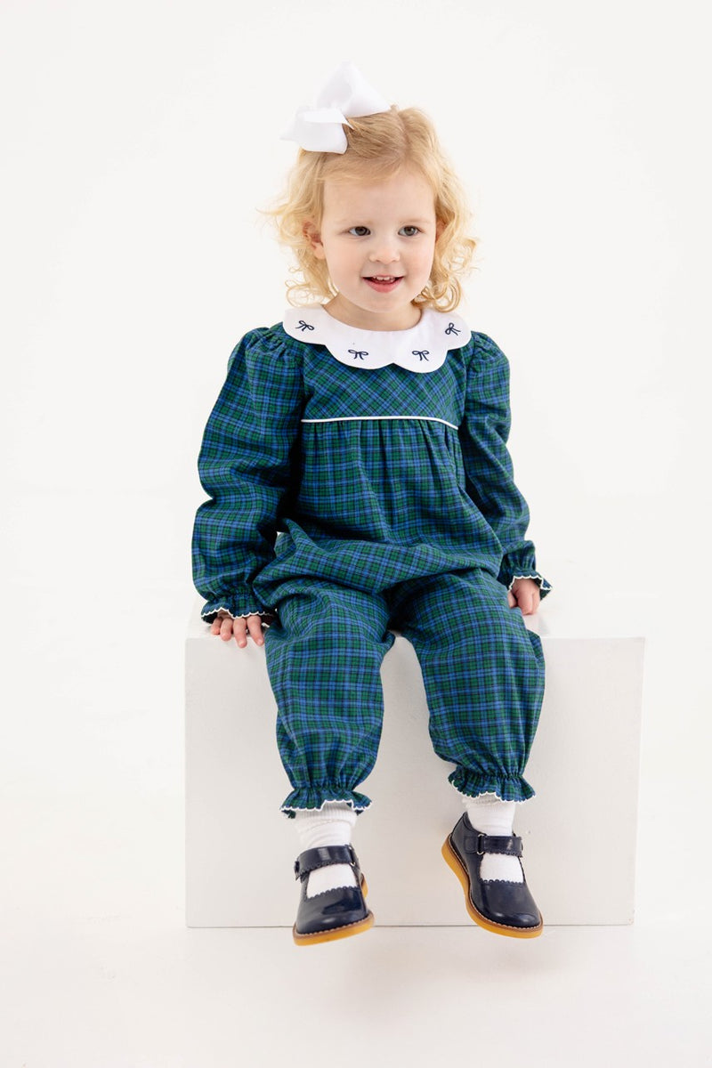 A young child with blonde hair and a white bow sits on a white block, wearing The Beaufort Bonnet Company’s Long Sleeve Britt Romper in Woodlake Point Plaid with Worth Avenue White—a charming autumn wardrobe addition—paired with a crisp white collar and black shoes.