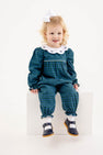 A young child with blonde hair and a white bow sits on a white block, wearing The Beaufort Bonnet Company’s Long Sleeve Britt Romper in Woodlake Point Plaid with Worth Avenue White—a charming autumn wardrobe addition—paired with a crisp white collar and black shoes.