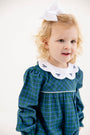 A young child with blonde curly hair, wearing a large white bow and the Long Sleeve Britt Romper in Woodlake Point Plaid with Worth Avenue White by The Beaufort Bonnet Company—featuring a white collar adorned with small navy bows—stands against a plain, light background.