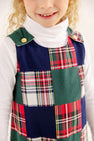 A child wears a Mercer Island Madras Juliet Jumper in a patchwork of solid green, navy blue, and colorful plaid fabric, styled with gold buttons at the shoulders over a white turtleneck.
