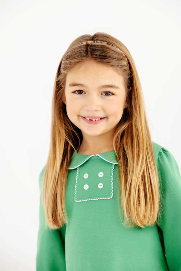 A young girl smiles wearing a The Beaufort Bonnet Company Long Sleeve Banks Button Dress - Kiawah Kelly Green with Worth Avenue White, featuring a white-trimmed collar and a decorative square panel with four white buttons.