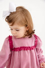 A young child with light brown hair and a white bow looks down, wearing The Beaufort Bonnet Company’s Debbie Day Bubble in Begley Berry Tweed—a charming pink outfit with darker pink ruffle trim—styled as an adorable baby girl ensemble against a plain white background.