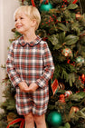 A young boy smiles in a John Hall Jon Jon - Paris Pike Plaid with Nantucket Navy long-sleeved romper with a rounded collar, posing in front of a decorated Christmas tree.