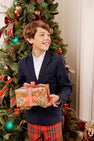 A boy wearing a Grandccasion™ Booker Blazer - Nantucket Navy with Tortoise Shell Buttons and plaid trousers holds a wrapped Christmas gift in front of a decorated tree.