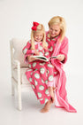 A woman and a young girl wear matching Payton Pajama Set - Distinguished Doggies with Hamptons Hot Pink by The Beaufort Bonnet Company while sitting in a white chair and reading a book together.