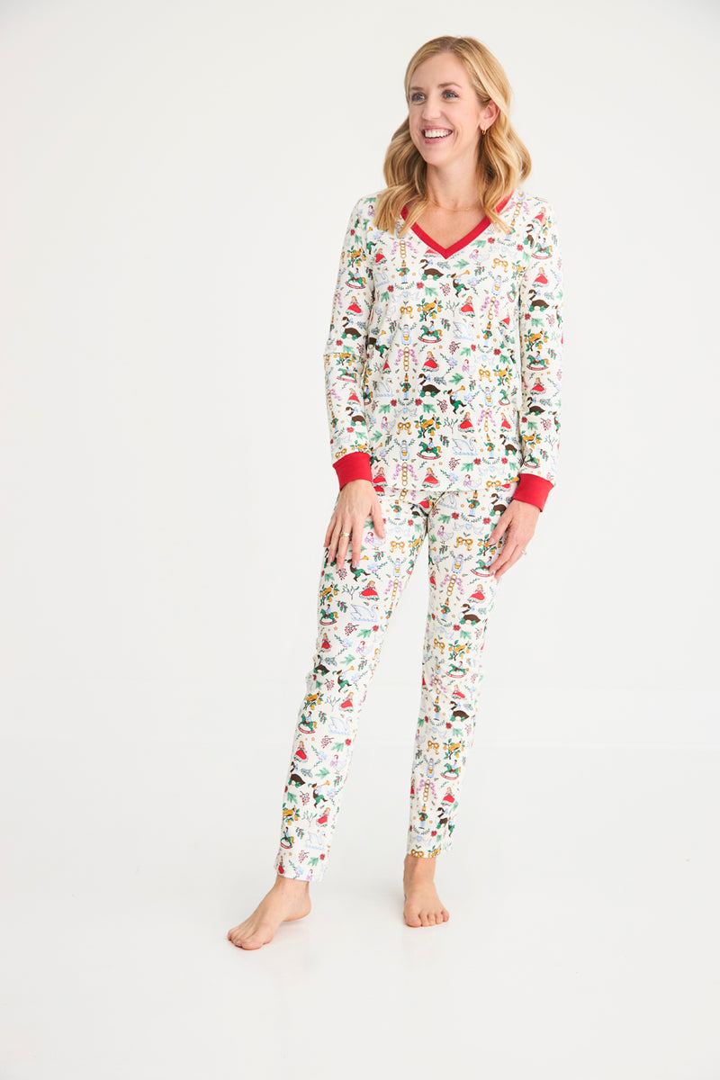 A woman stands barefoot on a white background, smiling as she wears The Beaufort Bonnet Company’s Coastal Living Lounge Set - 12 Days of Christmas with Richmond Red, crafted from soft Pima Cotton pajama fabric and adorned with colorful holiday-themed patterns and red trim.