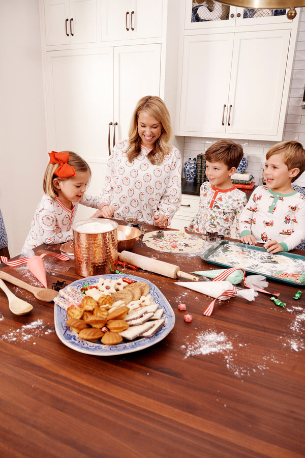 A woman and three children, all wearing The Beaufort Bonnet Company’s Snooze Button Set - North Pole Portrait with Richmond Red Stripe matching family pajamas, decorate cookies in a kitchen, surrounded by cookies, candy canes, and baking tools spread out on the wooden counter.