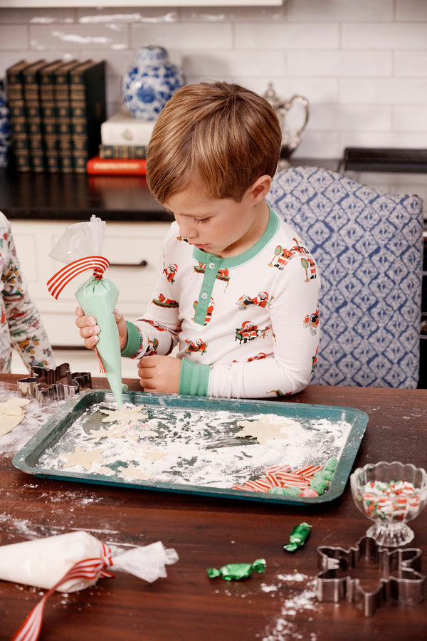 Wearing the Sutton’s Sweet Dream Set - Serious Santa Business by The Beaufort Bonnet Company, a young child uses a piping bag to decorate cookies at a kitchen table filled with baking supplies, cookie cutters, and candies.