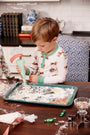 Wearing the Sutton’s Sweet Dream Set - Serious Santa Business by The Beaufort Bonnet Company, a young child uses a piping bag to decorate cookies at a kitchen table filled with baking supplies, cookie cutters, and candies.