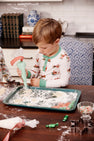 Wearing the Sutton’s Sweet Dream Set - Serious Santa Business by The Beaufort Bonnet Company, a young child uses a piping bag to decorate cookies at a kitchen table filled with baking supplies, cookie cutters, and candies.