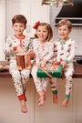 Three young children in Sutton’s Sweet Dream Set – Serious Santa Business pajamas from The Beaufort Bonnet Company sit on a kitchen counter, smiling. One holds a copper pot, another a green gift box with ribbon. Holiday treats are visible nearby, and the setting is bright and cheerful.