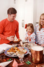 A family in Richmond Red holiday pajamas bakes cookies together at a kitchen table covered with decorated cookies, sprinkles, and baking tools. The dad wears The Beaufort Bonnet Company’s Men’s Long Sleeve Carter Crewneck in Richmond Red with Worth Avenue White Stork as they share laughter and holiday cheer.