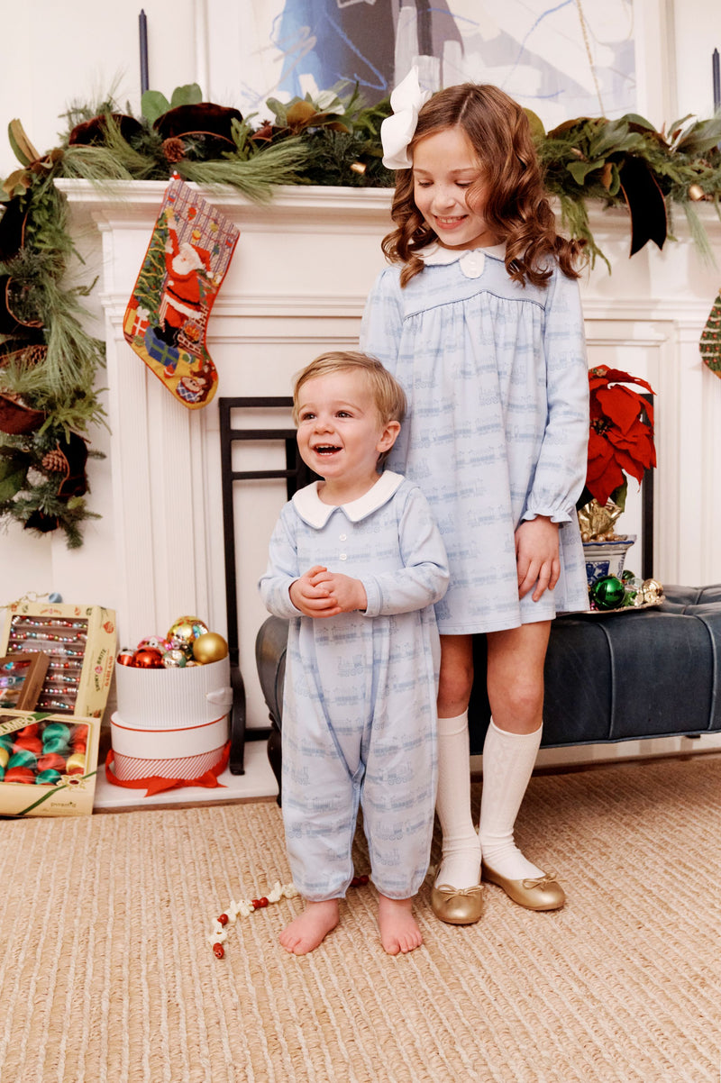 Two young children in matching blue pajamas stand before a festive fireplace adorned with garland, stockings, wrapped presents, and ornaments. The older girl beams in her The Beaufort Bonnet Company Long Sleeve Mary Dal Dress - Blue Choo Choos with Worth Avenue White, while the younger boy laughs, holding a red and white bead garland.
