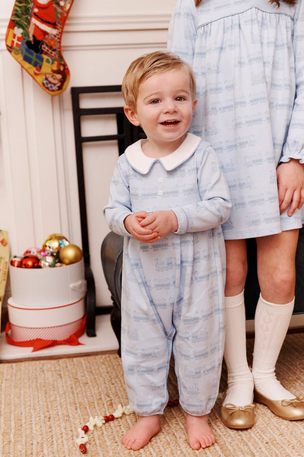 A young boy in a light blue Long Sleeve Bradford Romper - Blue Choo Choos with Worth Avenue White by The Beaufort Bonnet Company stands smiling next to an older girl in a matching dress, both posed in front of a fireplace adorned with Christmas stockings and ornaments.