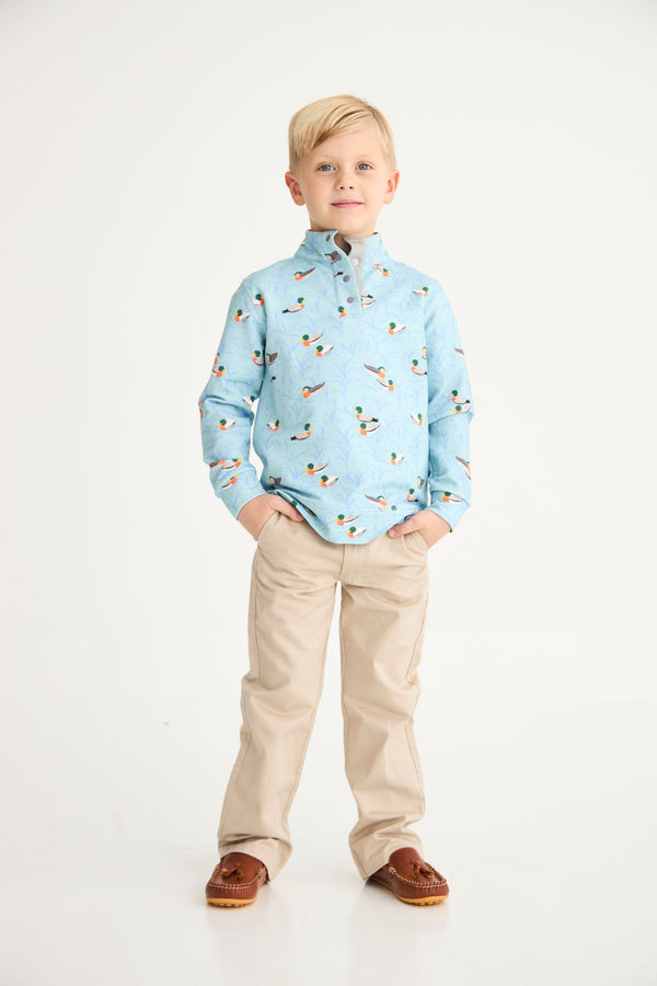 A young boy with blond hair stands with his hands in his pockets, wearing The Beaufort Bonnet Company’s Pendleton’s Popped Collar - Fine Weather For Ducks with Grantley Gray polo shirt, paired with beige pants and brown shoes, against a plain white background.