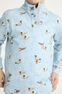 A child models The Beaufort Bonnet Company’s “Pendleton’s Popped Collar - Fine Weather For Ducks with Grantley Gray,” a light blue boys polo shirt adorned with mallard duck illustrations in flight and on the water. The shirt features a buttoned collar; the child’s face is partially out of frame.