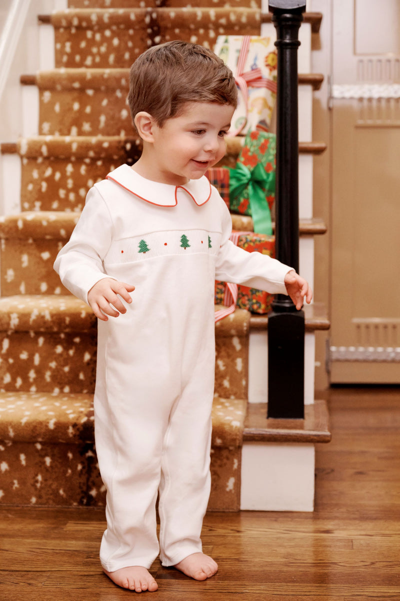 A toddler wearing a Samuel Smocked Romper - Worth Avenue White & Richmond Red with Trees stands at the bottom of a staircase.