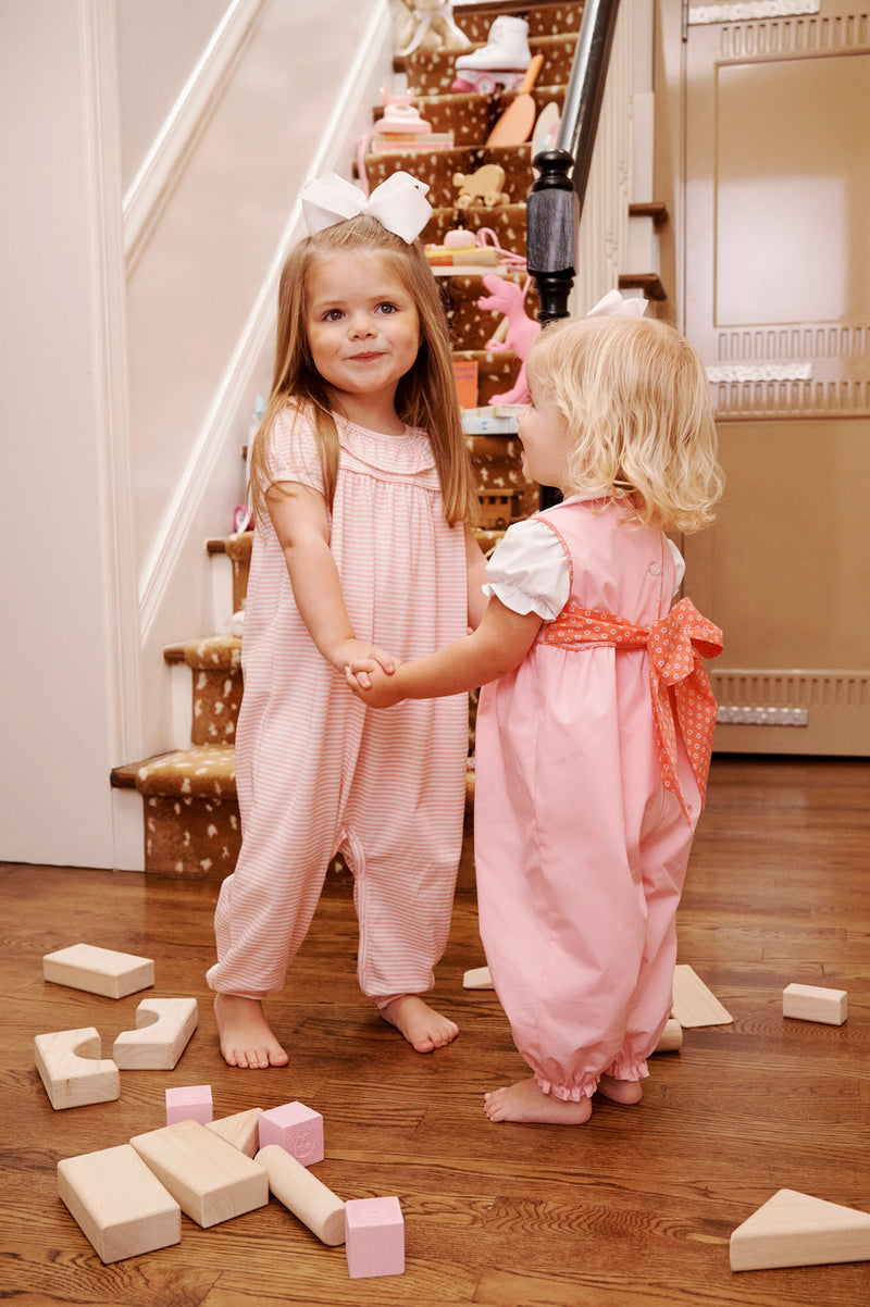 Two young girls wearing The Beaufort Bonnet Company’s Short Sleeve Ramona Ruffle Romper in Sandpearl Pink Stripe stand holding hands and smiling, surrounded by wooden blocks on the floor. A staircase with animal-print carpet and toys is visible in the background.