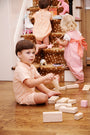 Three young children play indoors; one boy sits on the floor building with wooden blocks, while two girls wearing The Beaufort Bonnet Company’s Sir Proper’s Romper in Seashore Sherbet Stripe with Multicolor Stork enjoy playing with toys on and near carpeted stairs.