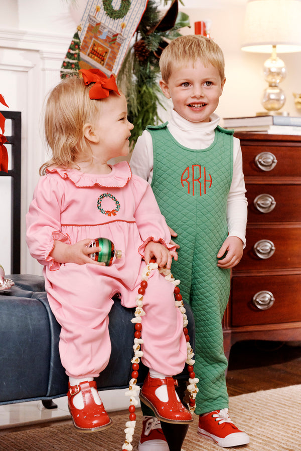 Two young children in festive holiday outfits smile indoors. The girl wears The Beaufort Bonnet Company’s Long Sleeve Ramona Ruffle Romper in Hamptons Hot Pink with Wreath Embroidery and holds a garland, accented by a red bow; the boy is dressed in a green quilted outfit with a monogram. Holiday decorations are visible in the background.