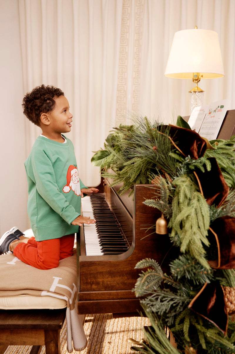 A young boy in The Beaufort Bonnet Company’s Cassidy Comfy Crewneck - Kiawah Kelly Green with Santa kneels on a piano bench, playing a piano adorned with festive greenery. A lit lamp and sheet music sit atop the piano, while a curtain drapes the background—creating a perfect scene for Christmas gatherings.