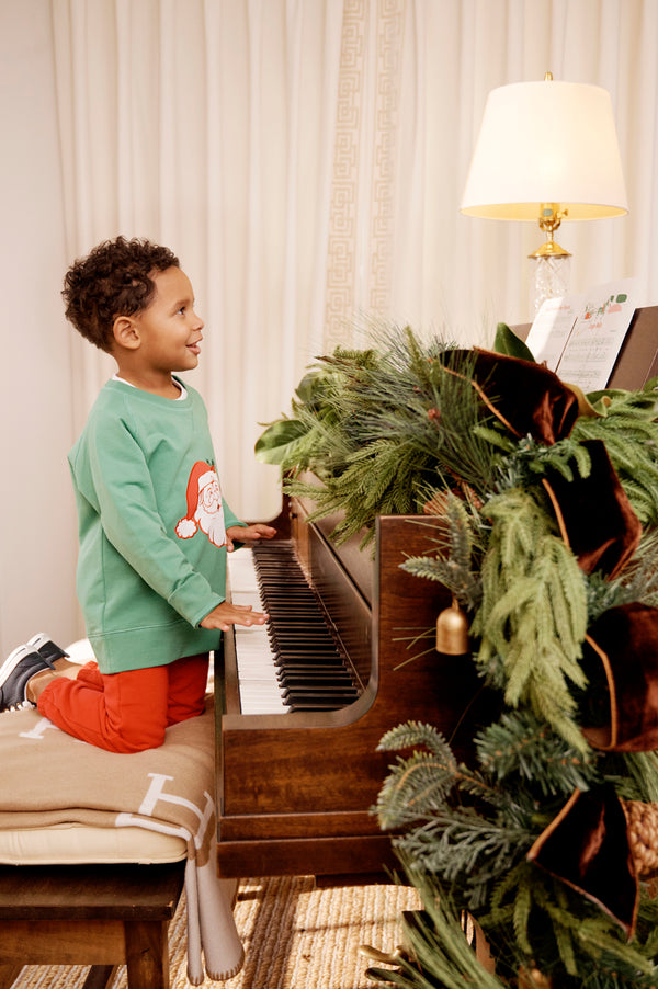 A young boy in The Beaufort Bonnet Company’s Cassidy Comfy Crewneck - Kiawah Kelly Green with Santa kneels on a piano bench, playing a piano adorned with festive greenery. A lit lamp and sheet music sit atop the piano, while a curtain drapes the background—creating a perfect scene for Christmas gatherings.