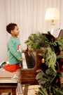 A young boy in The Beaufort Bonnet Company’s Cassidy Comfy Crewneck - Kiawah Kelly Green with Santa kneels on a piano bench, playing a piano adorned with festive greenery. A lit lamp and sheet music sit atop the piano, while a curtain drapes the background—creating a perfect scene for Christmas gatherings.