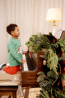 A young boy in The Beaufort Bonnet Company’s Cassidy Comfy Crewneck - Kiawah Kelly Green with Santa kneels on a piano bench, playing a piano adorned with festive greenery. A lit lamp and sheet music sit atop the piano, while a curtain drapes the background—creating a perfect scene for Christmas gatherings.