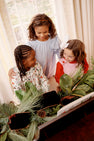 Three young girls stand together smiling, looking at a piano decorated with pine branches and pinecones near a window with cream curtains. One of the girls is wearing a festive Christmas dress, while another is dressed in The Beaufort Bonnet Company’s Juliet Jumper in Pier Party Pink with Richmond Red Bow.