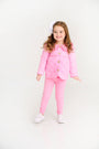 A young girl smiling while wearing a quilted pink jacket, Mitzy Sue Slacks (2-Pack) - Hamptons Hot Pink, and white sneakers.