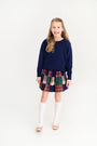 A young girl wears a polished outfit with a navy blue sweater, plaid skirt, gold flats, and Jefferies High Class Knee Socks - White from The Beaufort Bonnet Company.