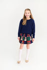 A young girl wears a polished outfit with a navy blue sweater, plaid skirt, gold flats, and Jefferies High Class Knee Socks - White from The Beaufort Bonnet Company.