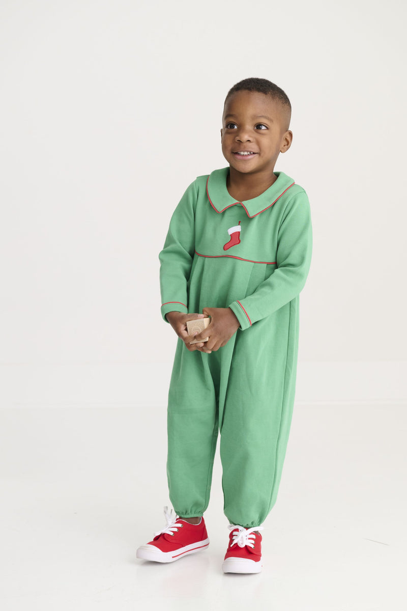 A young child in The Beaufort Bonnet Company’s Long Sleeve Bradford Romper – Kiawah Kelly Green with Stocking Embroidery stands smiling, wearing red sneakers and holding something in their hands against a plain white background.