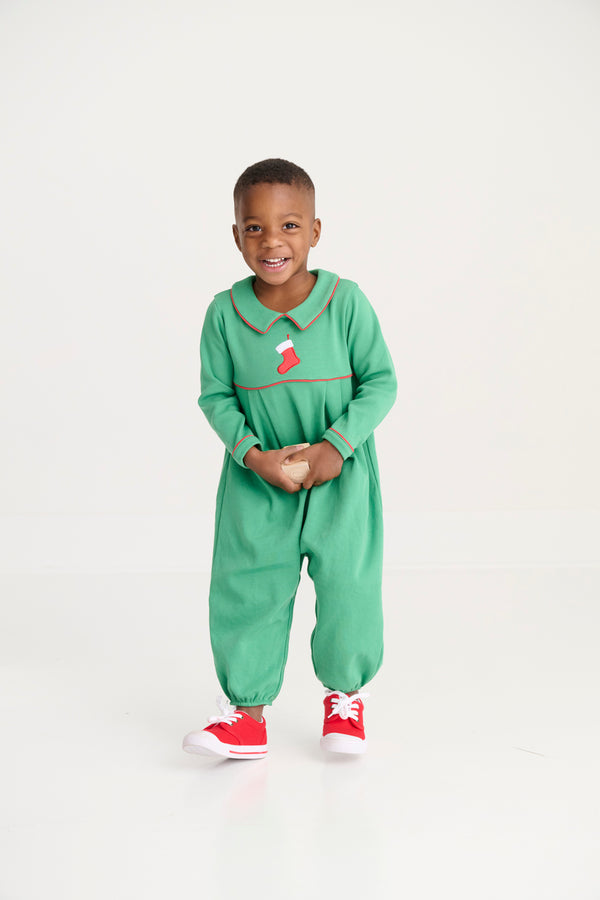 A young child smiles while standing in The Beaufort Bonnet Company’s Long Sleeve Bradford Romper - Kiawah Kelly Green with Stocking Embroidery, paired with red sneakers, against a plain white background.