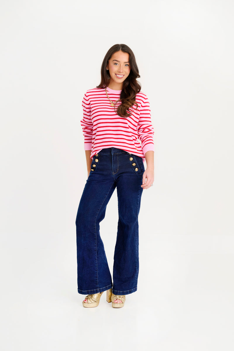 A woman stands smiling on a plain white background, wearing The Beaufort Bonnet Company’s Women’s Cissy Sweater in Pier Party Pink & Richmond Red Stripe. She pairs the festive cotton sweater with dark blue high-waisted wide-leg jeans featuring gold buttons and finishes her look with gold shoes.