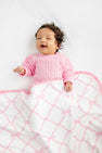 A smiling baby with curly hair wearing a pink sweater lies on a white surface, partially covered by The Beaufort Bonnet Company’s Baby Buggy Blanket in the Belle Meade Bow with Pier Party Pink design—a charming white blanket featuring a pink pattern and ruffled edges—making this scene perfect for anyone searching for an ideal baby shower gift.