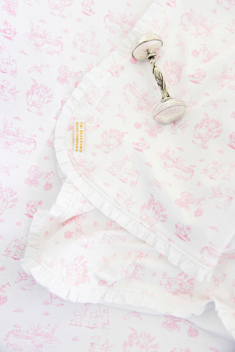 A silver rattle rests on Sir Proper’s Sweet Arrival (Hamptons Hot Pink) Accessories Gift Bundle from The Beaufort Bonnet Company, featuring a Pima cotton baby buggy blanket with pink illustrations and ruffled edges. A yellow tag displaying THE BEAUFORT BONNET COMPANY is visible on the blanket, making this charming bundle—including a matching burp cloth and bib—a delightful choice for baby shower gifts.