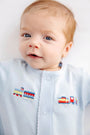 A baby with light hair and blue eyes wears The Beaufort Bonnet Company’s Twinkle Twinkle 2-in-1 - Buckhead Blue with Trains romper, which features an embroidered colorful toy train on the front. The baby looks at the camera with a slight smile.