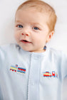 A baby with light hair and blue eyes wears The Beaufort Bonnet Company’s Twinkle Twinkle 2-in-1 - Buckhead Blue with Trains romper, which features an embroidered colorful toy train on the front. The baby looks at the camera with a slight smile.
