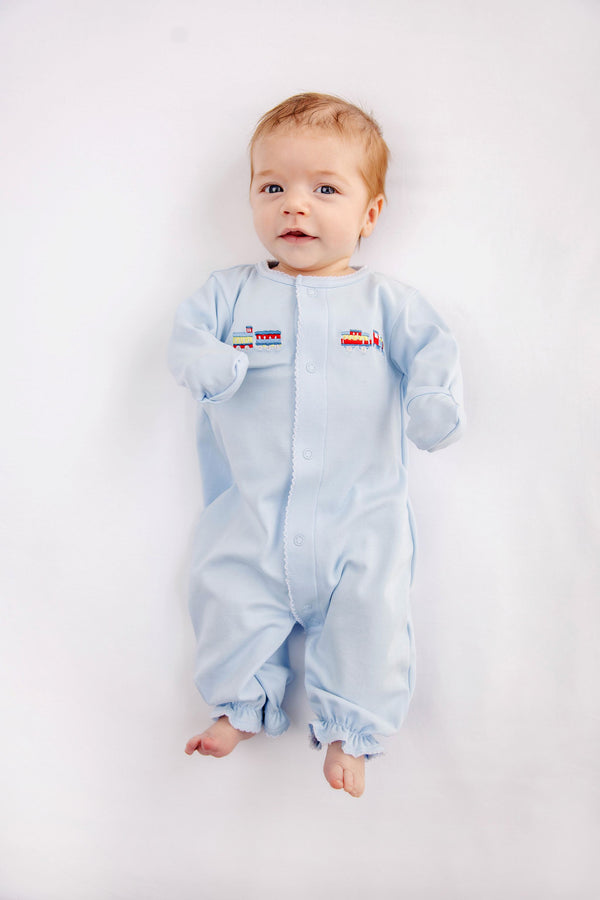 A baby with light hair and blue eyes lies on a white surface, wearing The Beaufort Bonnet Company’s Twinkle Twinkle 2-in-1 - Buckhead Blue with Trains romper in soft Pima cotton, featuring small embroidered train designs on the chest. The baby is looking up and smiling slightly.
